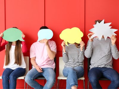 Četri cilvēki sēž pie sarkanas sienas, aizklājot savas sejas ar dažādu krāsu runas un domu burbuļiem – zaļu, gaiši zilu, dzeltenu un baltu.