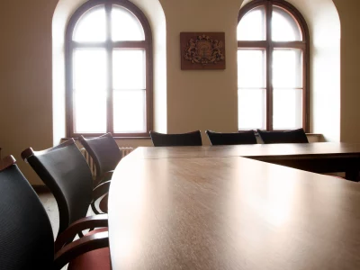 Tukša sanāksmju telpa ar garu koka galdu un krēsliem. Divi gaiši arkveida logi apgaismo telpu, un starp tiem pie sienas ir koka emblēma.Kuldīgas novada pašvaldības mazā zāle.