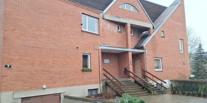 Sarkana ķieģeļu ēka ar stāviem jumtiem un dažādu formu logiem. Galvenā ieeja ar koka margām ved uz durvīm zem nelielas nojumes.