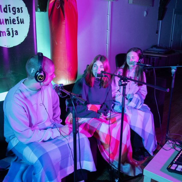 Trīs jaunieši, divas sievietes un vīrietis, sēž zem segām, runā mikrofonos un valkā austiņas, ierakstot podkāstu purpursarkanās un zilās gaismās. Fonā redzams plakāts ar uzrakstu "Kuldīgas Jauniešu māja".