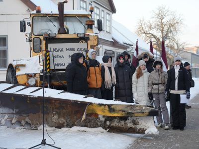 Grupa cilvēku, tostarp jaunieši, ziemas apģērbā stāv sniegotā laukumā pie liela dzeltena sniega tīrītāja ar uzgriezni priekšā. Daži no viņiem tur Latvijas karogus. Uz sniega tīrītāja ir zīme ar tekstu "LABĀK MANU GALVU ŅĒMA N MANU".