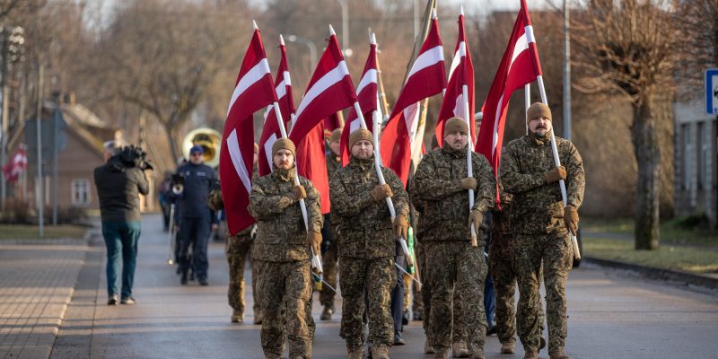 Vairāki vīrieši militārās maskēšanās formās soļo pa ielu, nesot paceltus Latvijas karogus. Fonā redzams orķestris