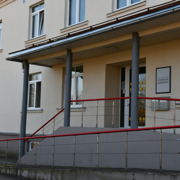 Gaišas krāsas ēka ar uzbrauktuvi un sarkaniem margām pie ieejas, virs kuras ir zīme ar uzrakstu "SOCIĀLAIS DIENESTS". Pa kreisi plīvo Latvijas karogs.