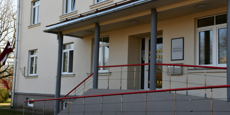 Gaišas krāsas ēka ar uzbrauktuvi un sarkaniem margām pie ieejas, virs kuras ir zīme ar uzrakstu "SOCIĀLAIS DIENESTS". Pa kreisi plīvo Latvijas karogs.