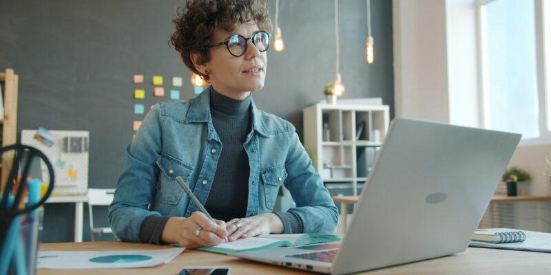 Pārdomās iegrimusi sieviete ar lokainiem matiem un brillēm sēž pie galda, rakstot piezīmju grāmatiņā ar pildspalvu, kamēr priekšā atvērts portatīvais dators modernā birojā.