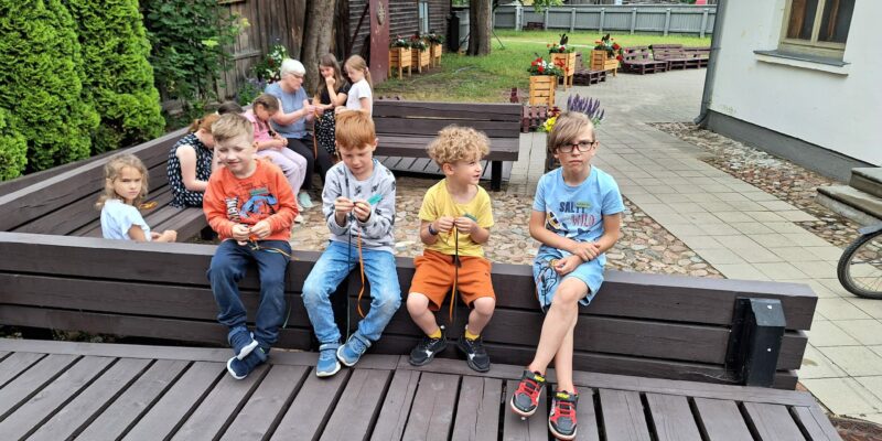 Āra nodarbībā bērnu un pieaugušo grupa veido rokdarbus. Priekšplānā četri zēni sēž uz koka terases, divi no viņiem aktīvi pin krāsainas auklas. Zēnam labajā pusē ir gaiši zils T-krekls ar uzrakstu "SALTY WILD". Fonā redzamas citas personas, vecas koka ēkas un koki.