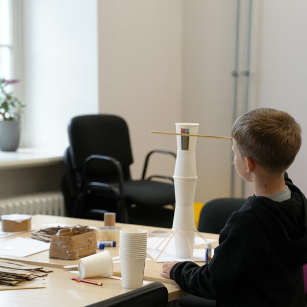 Zēns no muguras stāv pie galda un skatās uz no baltām papīra krūzītēm un koka nūjiņām izveidotu torni. Uz galda redzami citi rokdarbu materiāli.