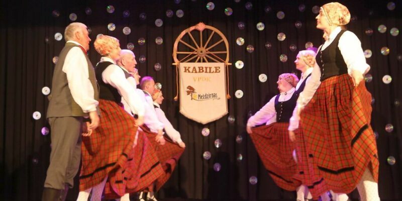 Pieaugušo tautas deju kolektīvs tradicionālos tērpos dejo uz skatuves. Dejotāji, vīrieši pelēkos uzvalkos un sievietes sarkanos rūtainos svārkos ar melnām vestēm, ir izvietoti divās rindās, sievietēm turot paceltus svārkus. Fonā tumšs aizkars ar izkārtotiem kompaktdiskiem un baneris, uz kura redzams uzraksts "KABILE VPDK Meždzirnas" un stilizēts koka rats.