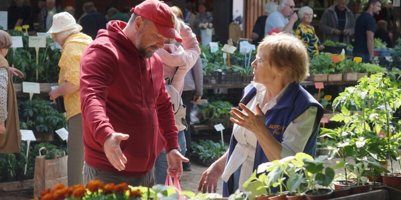 Vīrietis sarkanā džemperī un sieviete zilā vestē ar uzrakstu "AUTO F" sarunājas tirgū, ko ieskauj daudz podos audzētu stādu.